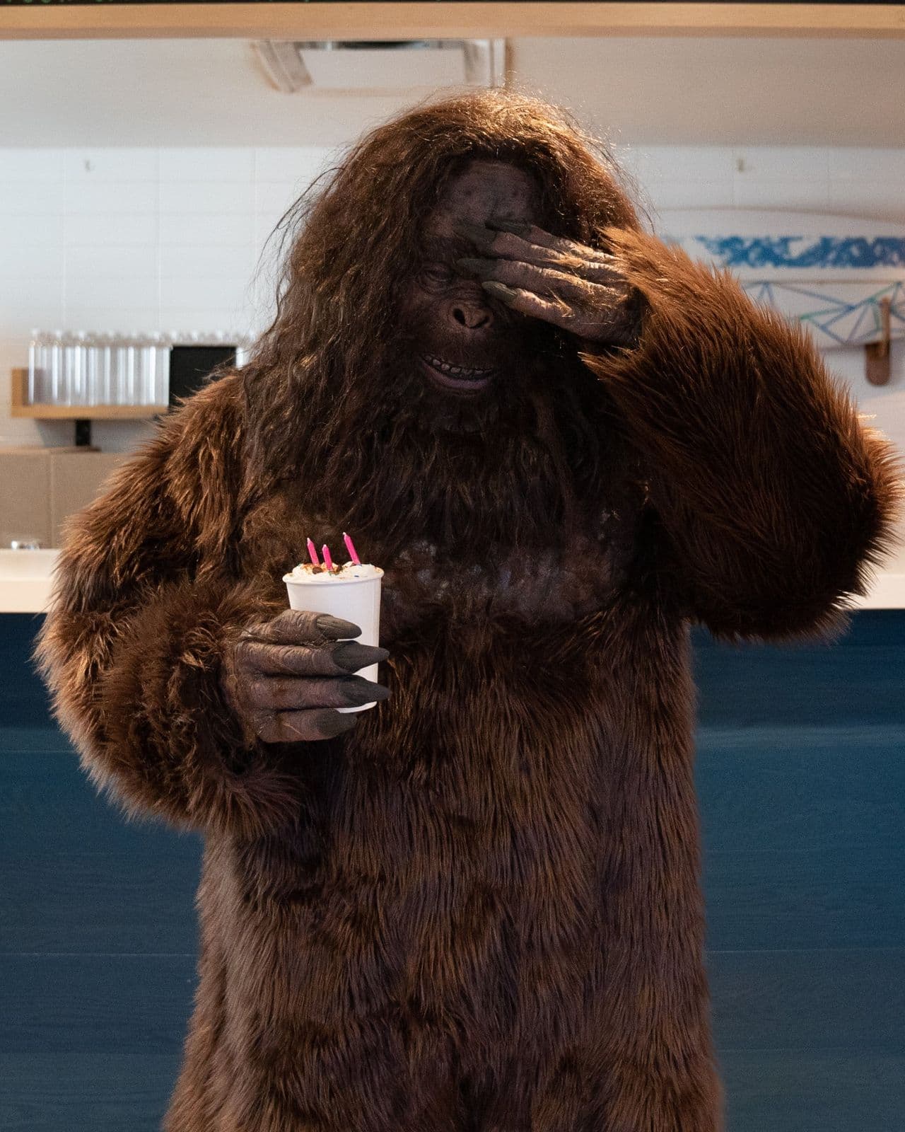 Bigfoot covering his face, holding a sprinkle-topped drink with birthday candles at a coffee counter